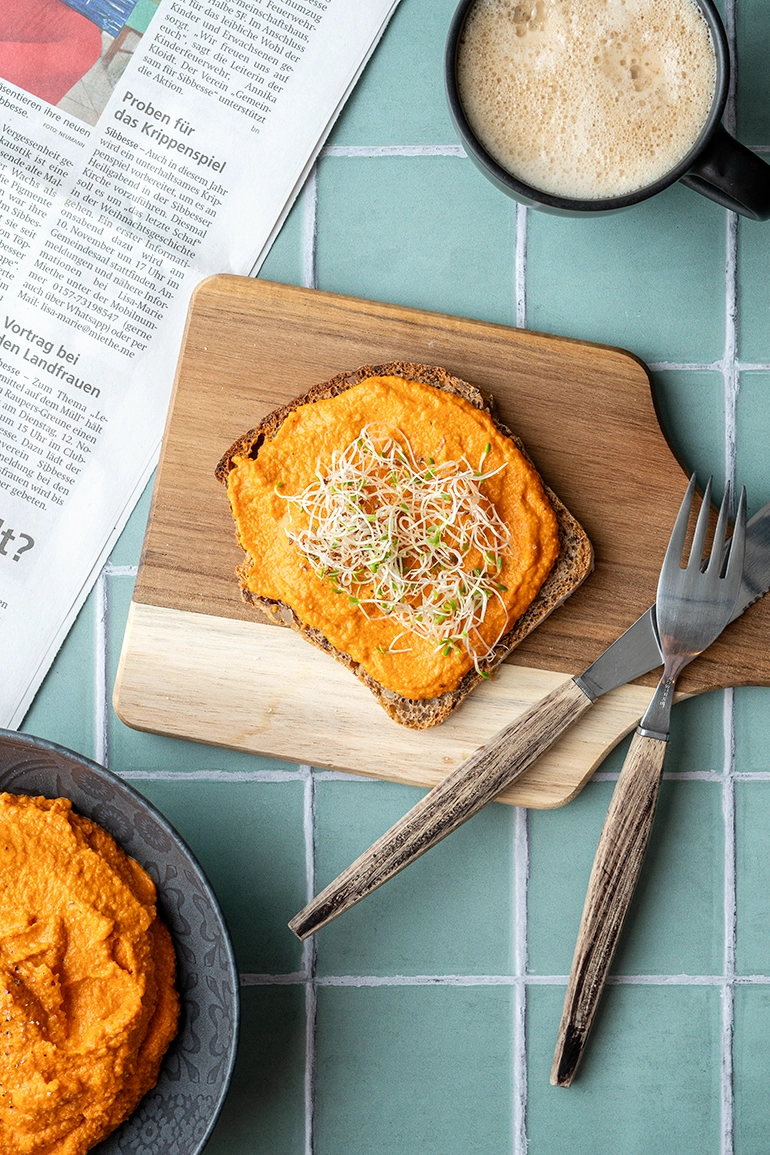 vegan_rezept_einfacher_Cashew_Paprika_Dip_3 Eine Scheibe Brot mit Cashew-Aufstrich auf einem Brett mit Messer und Gabel