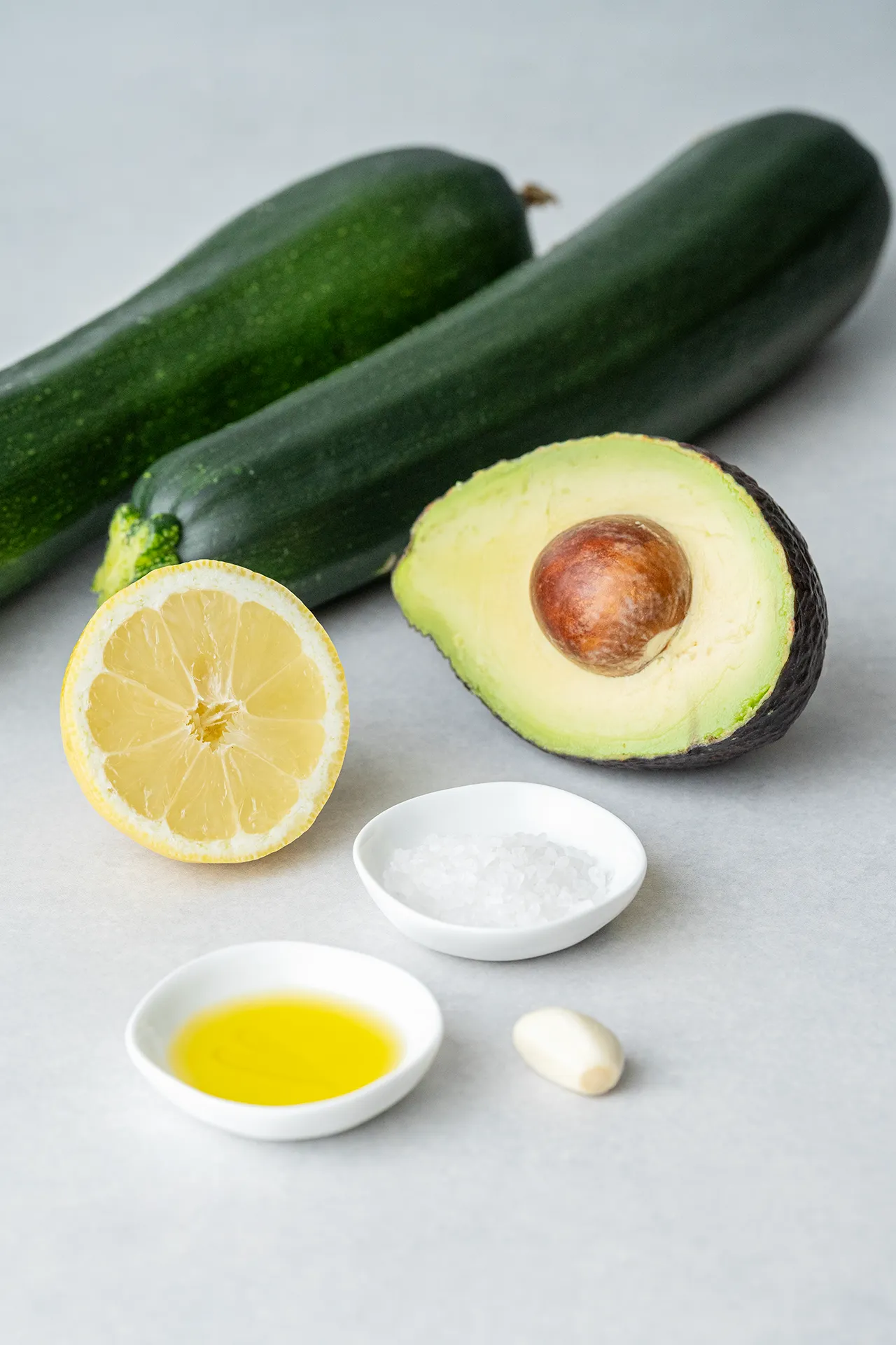 Zucchinis, Avocado, Zitrone, Knoblauch, Salz und Olivenöl auf hellem Untergrund