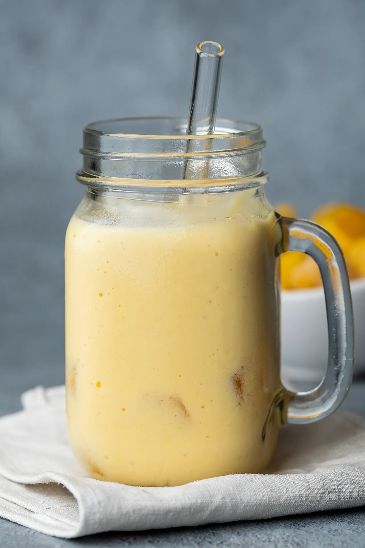 Veganer Mango Lassi – einfaches Rezept mit nur 4 Zutaten 2 Veganer Mango Lassi im Glas auf einem Tuch dahinter Mangostücke in einer Schale